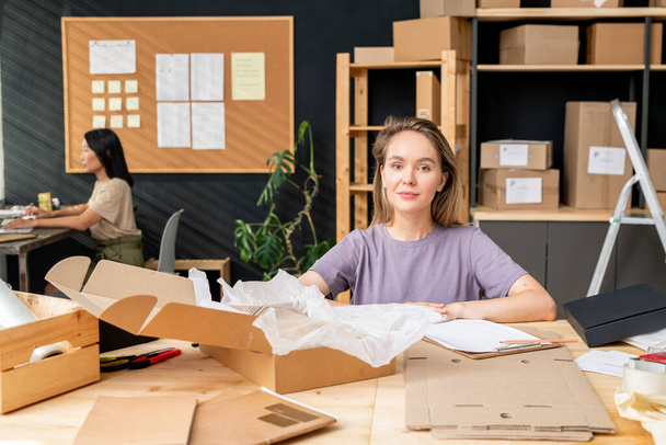 Young pretty female worker of online shop office sitting by table in front of camera, packing orders of clients and checking data in document - Zdjęcie, obraz