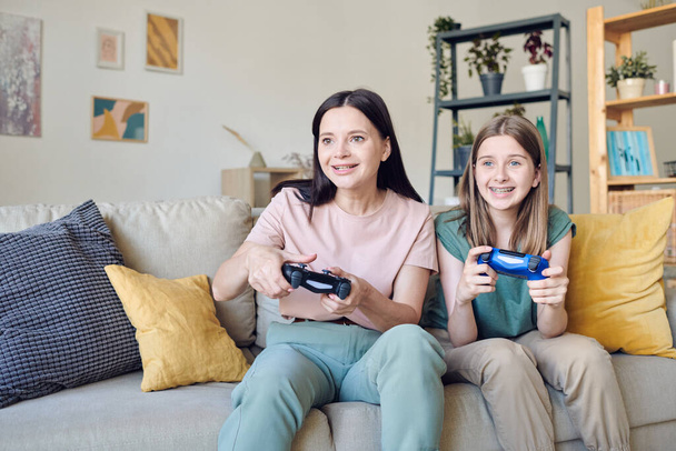 Young smiling mother and cute teenage girl with joysticks pressing buttons during video game while sitting on couch against shelves by wall - 写真・画像