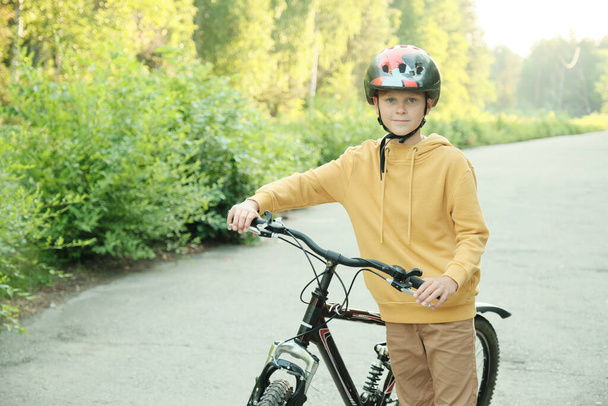 Jeune garçon en sweat à capuche jaune, pantalon beige et casque de protection debout avec vélo sur la route devant la caméra contre les arbres verts - Photo, image