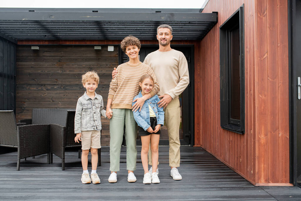 Щаслива родина молодих батьків і двох маленьких дітей, що стоять на дерев'яній підлозі патіо свого нового будинку з чорними кріслами позаду
 - Фото, зображення