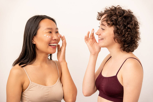 Two young laughing intercultural females in tanktops applying rejuvenating cream on undereye area while looking at one another in isolation - Photo, Image