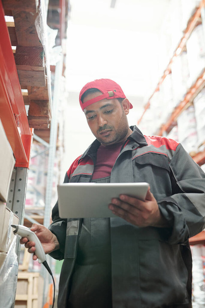 Joven trabajador de raza mixta serio de almacén usando tableta mientras está parado entre estantes con mercancías embaladas y códigos qr de escaneo - Foto, imagen