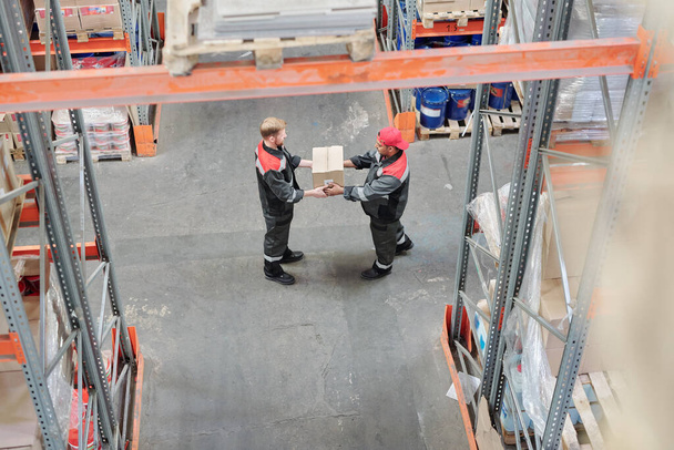 Visão geral do jovem trabalhador do armazém que passa caixa embalada para seu colega, enquanto ambos de pé pelo corredor entre racks carregados - Foto, Imagem