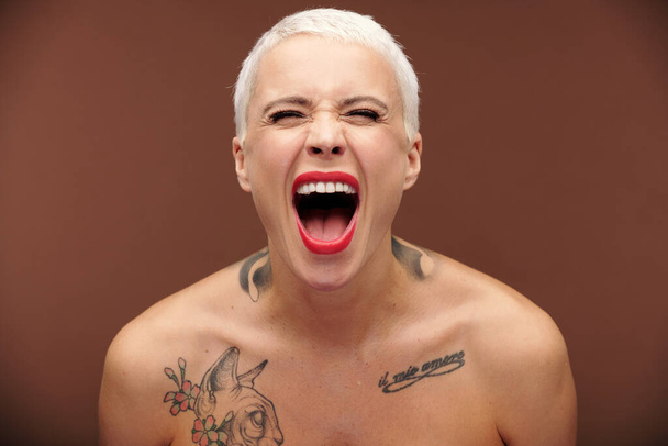 Giovane femmina maschile con capelli biondi corti, rossetto rosso sulle labbra e tatuaggi sul petto e sul collo urlando ed esprimendo aggressività - Foto, immagini