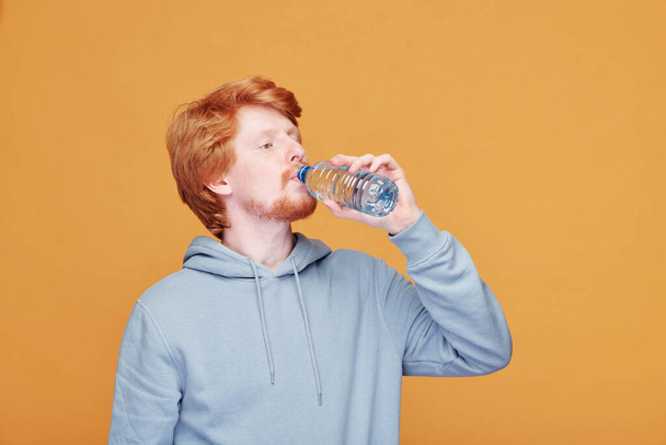 Portrait of smiling handsome young redhead man in hoodie applying toothpaste on toothbrush against orange background - Photo, Image