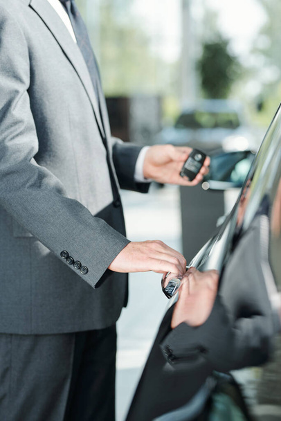 Jeune vendeur élégant de voitures neuves en costume en utilisant le système d'alarme à télécommande tout en se tenant près de l'une des automobiles et va ouvrir la porte - Photo, image