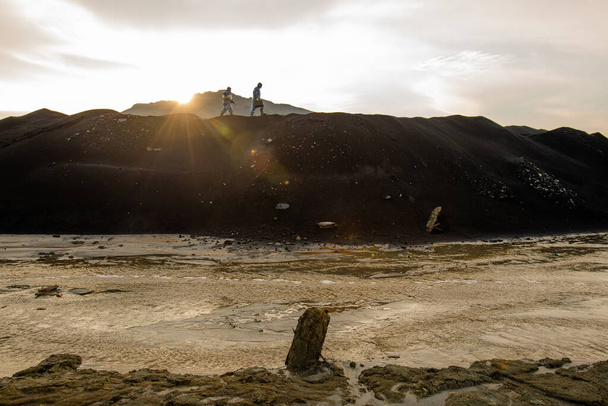 Petites silhouettes de deux chercheurs contemporains en combinaison de protection se déplaçant le long de la colline contre le ciel gris nuageux au coucher du soleil - Photo, image