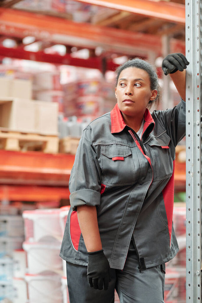 Работница среднего возраста смешанной расы с большим современным складом в перчатках и рабочей одежде смотрит в сторону во время отдыха на стойке - Фото, изображение