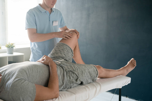 mannelijke patiënt liggend op medische bank terwijl jonge fysiotherapeut masseren zijn gebogen linkerknie tijdens revalidatie cursus - Foto, afbeelding