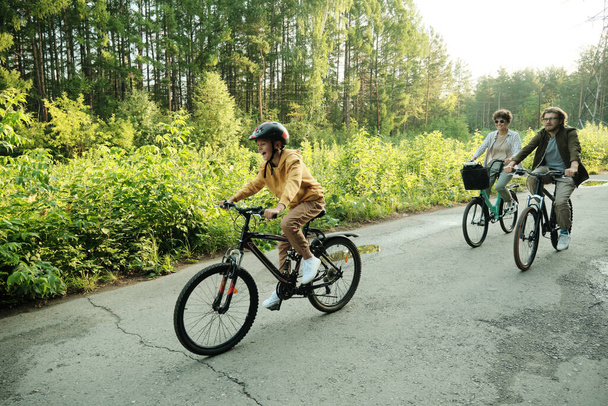Νεαρό ζευγάρι και ο γιος τους ιππασία ποδήλατα κατά μήκος του δρόμου σε φυσικό περιβάλλον κατά πράσινα δέντρα και θάμνους το Σαββατοκύριακο του καλοκαιριού - Φωτογραφία, εικόνα