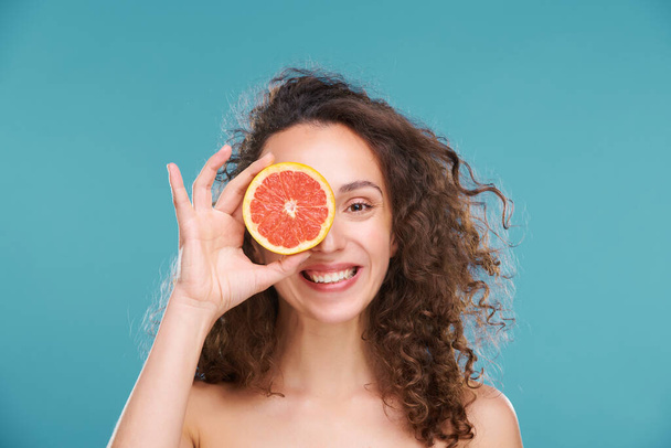 Junge, fröhliche Frau mit einem gesunden Lächeln und einer gesunden Haut, die dich mit dem linken Auge ansieht, während sie eine frische Grapefruitscheibe neben die andere hält - Foto, Bild