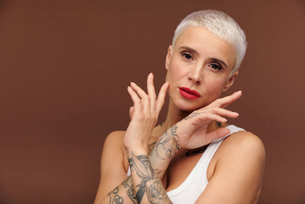 Mature blond female with tattooes on arms surrounding her face by hands and looking at you while standing in front of camera in isolation - Foto, immagini