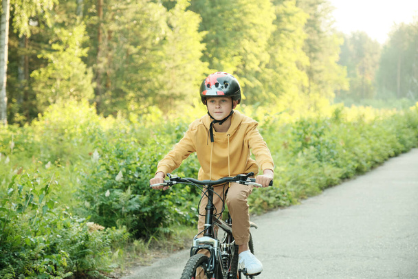 Estudante contemporâneo ativo em casualwear e capacete de proteção equitação nova bicicleta ao longo da estrada no parque no dia ensolarado ou de manhã no lazer - Foto, Imagem