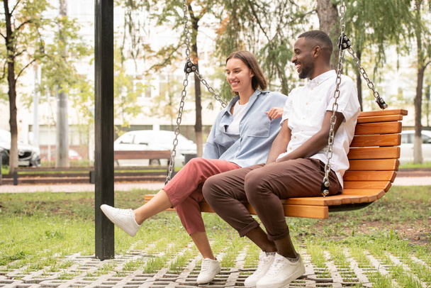 Szczęśliwa młoda para międzykulturowa w stroju casualwear siedzi na drewnianym huśtawce w parku lub na placu, rozmawiając i śmiejąc się podczas letniego dnia - Zdjęcie, obraz