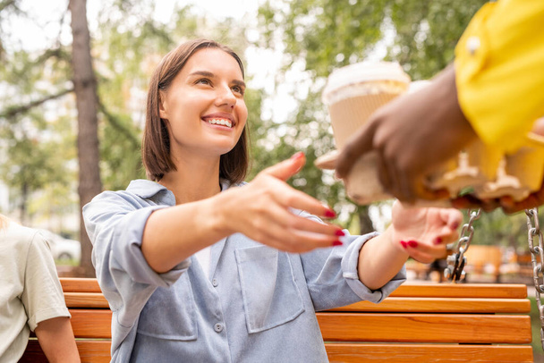 Hands of courier in yellow jacket passing two drinks from restarant or cafe to young smiling brunette woman sitting on wooden bench in park - Photo, Image