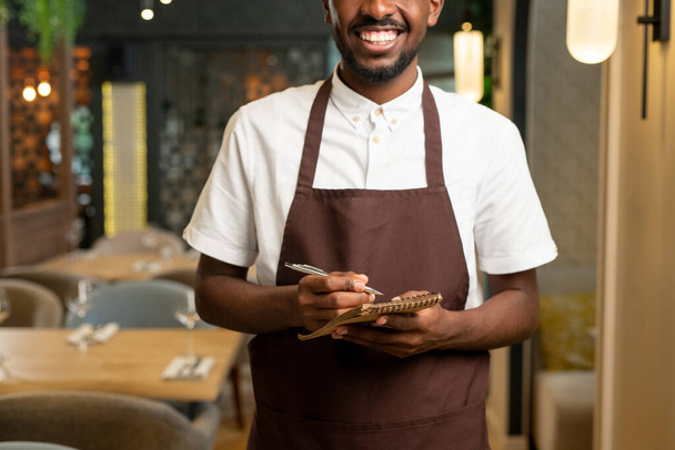Руки юного официанта с зубастой улыбкой, держащего ручку и блокнот, стоя перед камерой в ресторане и принимая заказы - Фото, изображение