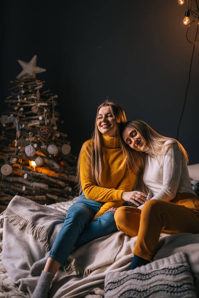 две девушки в свитерах сидят на диване в новогодней комнате, одна девушка кладет голову на плечо другой. - Фото, изображение