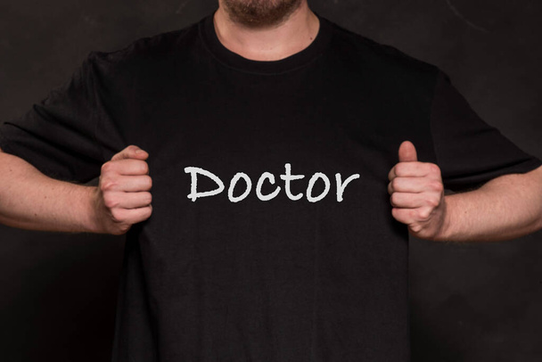 mężczyzna w czarnej koszuli z napisem DOCTOR, czarne tło - Zdjęcie, obraz