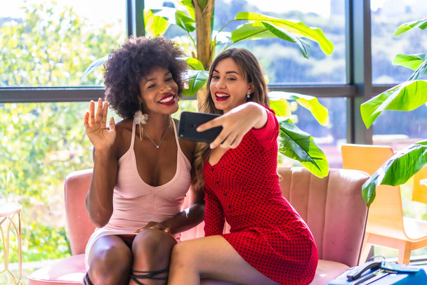 Lifestyle, two friends who have just arrived at the hotel with suitcases taking a selfie with the mobile, a Caucasian blonde girl and a black girl with afro hair, a pink dress and a red dress. - Photo, Image