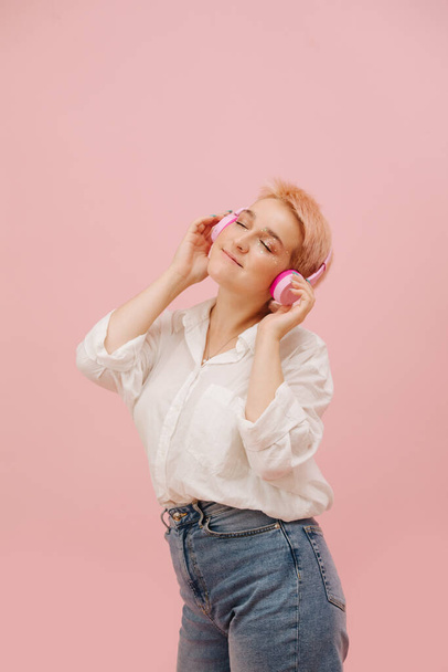 Immersed young woman with short hair enjoying music, listening to wireless headphones. Over pink background. She has short dyed blond hair and stars eye makeup. She wears mom jeans and dress shirt. - Zdjęcie, obraz
