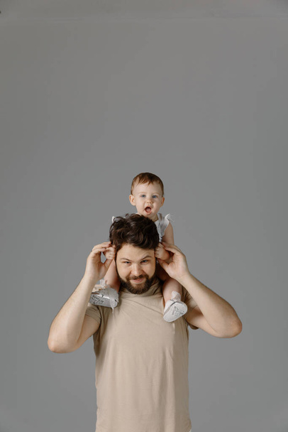 Father and daughter relations - smiling in studio, isolated on gray background. A young girl sits atop the Shoulders of her Dad, lifestyle - Foto, Bild