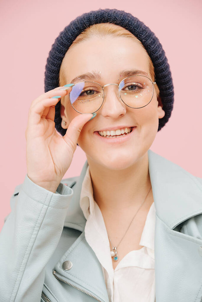 Eccentric young woman in a cap holding her round glasses, and openly smiling. Over pink background. Model has short hair and stars eye make up. - Fotó, kép