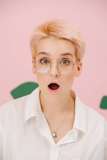 Surprised shocked young woman with short dyed blond hair and round glasses and stars eye makeup. Over pink background. Mouth opened wide. - Photo, Image