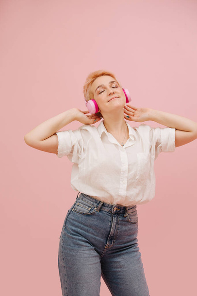 Dansing young woman with short hair enjoying music, listening to wireless headphones, swaying hips. Over pink background. She has short dyed blond hair and wears mom jeans. Her eyes closed in pleasure - Fotografie, Obrázek