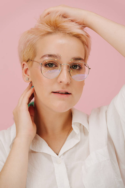 Portrait of a sensual young woman with short dyed blond hair and round glasses and stars eye makeup. Over pink background - Fotó, kép
