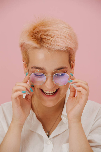 Portrait of a smiling young woman with short dyed blond hair and round glasses and stars eye makeup. She's laughing, looking down. Over pink background - Photo, Image