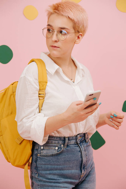Young woman with short dyed blond hair and round glasses. She wears mom jeans, dress shirt and yellow backpack. Holding smartphone. Over pink wall with green and yellow paper shapes glued to it. - Foto, Imagen