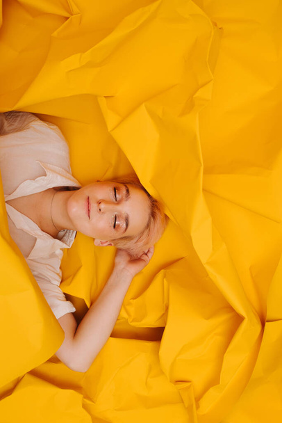 Happy satisfied young woman lying on her back on yellow cramped paper. She has short dyed blond hair and stars eye makeup. Her eyes closed. - Фото, изображение