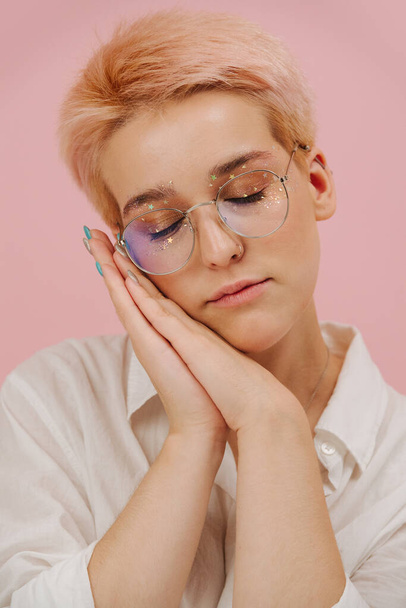 Young woman napping on her joined hands. She has short dyed blond hair and round glasses and stars eye makeup. Over pink background - Foto, Imagen