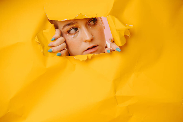 Young woman peeking through a hole in a big sheet of cramped yellow paper. Her hands on edges. She has short dyed blond hair and stars eye makeup. - Foto, Bild