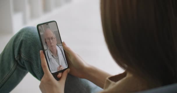 Arzthelferin trägt weißen Kittel, Kopfhörer-Video ruft entfernte Patientin auf dem Smartphone an. Arzt spricht mit Klient mittels virtueller Chat-Telefon-App. Telemedizin, Fernversorgung  - Filmmaterial, Video