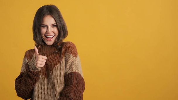 Vonzó vidám barna lány hangulatos kötött pulóver boldogan mutatja hüvelykujját felett színes háttér - Fotó, kép