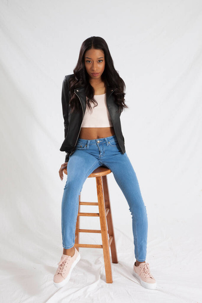 Hübsche schwarze Frau in Lederjacke und Jeans sitzt auf einem Holzhocker - Foto, Bild