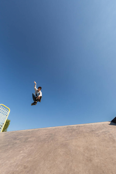 bruslař ve vzduchu po skoku na rampu dělat trik v bruslovém parku. mladý běloch, hnědý, s tričkem a džínama. slunečný den - Fotografie, Obrázek