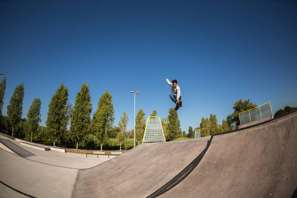 een rolschaatser in de lucht na het springen op een helling met een truc in een skatepark. Jonge blanke man, bruin, met t-shirt en jeans. zonnige dag - Foto, afbeelding