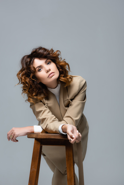 elegant woman in beige suit posing on wooden chair isolated on grey - Zdjęcie, obraz