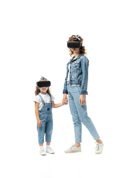 madre e hija con trajes de mezclilla y auriculares de realidad virtual tomados de las manos aisladas en blanco - Foto, Imagen