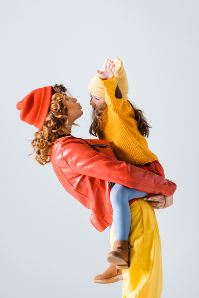 side view of mother in colorful red and yellow outfit holding and kissing daughter isolated on grey - Fotografie, Obrázek