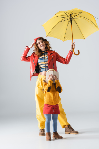 mother and daughter in colorful red and yellow outfits with umbrella and book on grey background - Фото, изображение