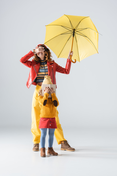 mother and daughter in colorful red and yellow outfits with umbrella and book covering faces on grey background - Photo, Image