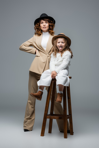 elegant mother posing near daughter showing tongue on chair on grey - Foto, immagini