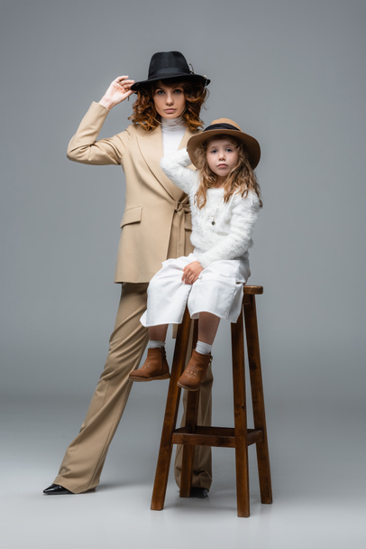 elegant mother and daughter in white and beige outfits posing on chair on grey - Foto, imagen