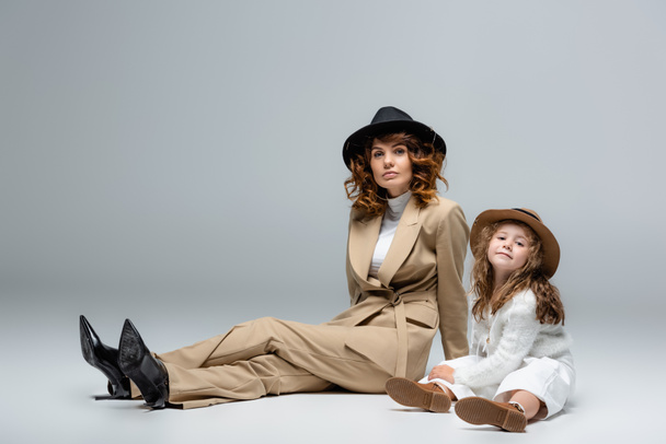 elegant mother and daughter in white and beige outfits and hats posing on floor on grey background - Fotografie, Obrázek