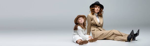 elegante madre e hija en trajes blancos y beige y sombreros posando en el suelo sobre fondo gris, plano panorámico - Foto, imagen