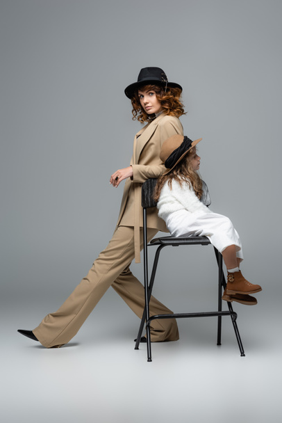 side view of elegant mother and daughter in white and beige outfits and hats posing on chair on grey background - Photo, Image