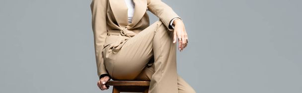 cropped view of elegant woman in beige suit posing on wooden chair isolated on grey, panoramic shot - 写真・画像